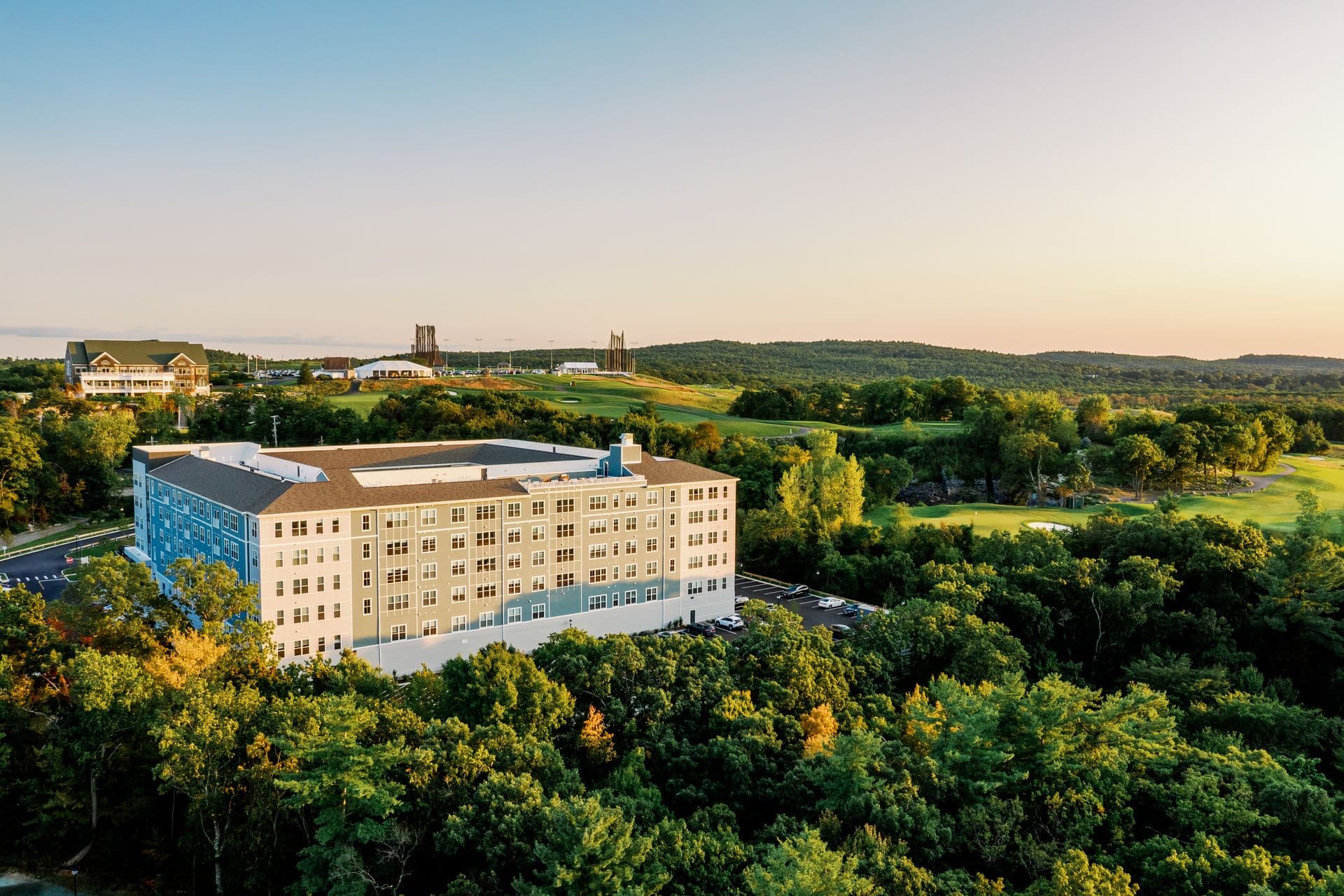 Golden-hour aerial of The Q in Quincy, MA, surrounded by tree canopy and fairways — landscape by Hynes